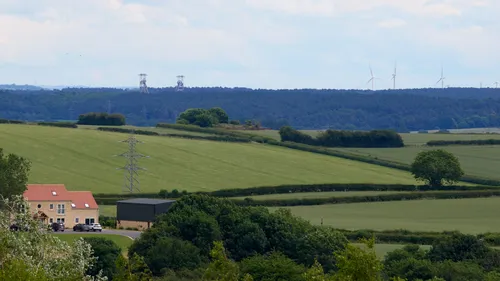 This landscape photograph captures a rural scene in late morning during the summer, showcasing a gently rolling expanse of agricultural fields. In the foreground, there is a farmhouse and outbuildings, surrounded by lush green hedgerows and open spaces typical of agriculture in the region. The main subjects of coal and wind are represented by the prominent mining headgear structures in the distance, which are framed by a forested area, and a row of wind turbines located on the horizon. Power lines cut across the fields, emphasizing the connection between traditional mining and modern renewable energy sources. The combination of mining infrastructure and wind turbines illustrates the transition between coal and wind energy within a landscape shaped by agriculture and power generation.
